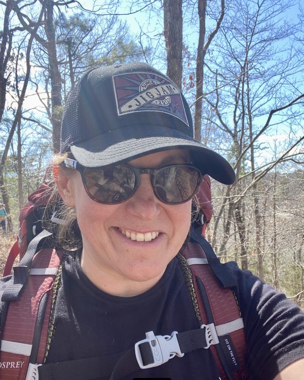 Conni wearing a baseball hat, and sunglasses with backpack smiling at the camera with the woods behind her. 